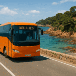Un autobús naranja de Rodalies circula por una carretera de la Costa Brava, con el panel LED delantero mostrando el operador. El paisaje combina mar turquesa, acantilados rocosos y pinares mediterráneos bajo un cielo despejado.