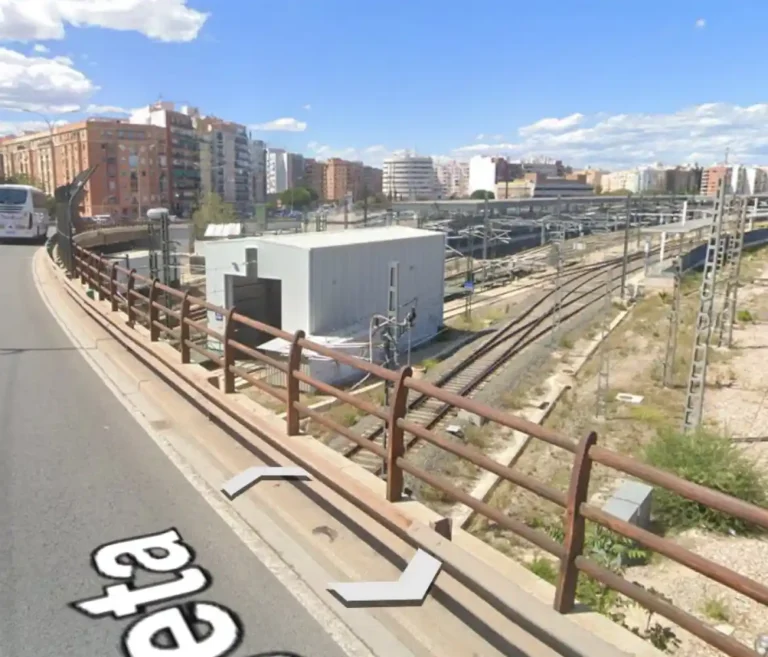 Vista desde un puente de las vías de tren y el cambiador de ancho en la salida de la estación Valencia-Joaquín Sorolla, con una caseta técnica gris y el perfil urbano de la ciudad al fondo.
