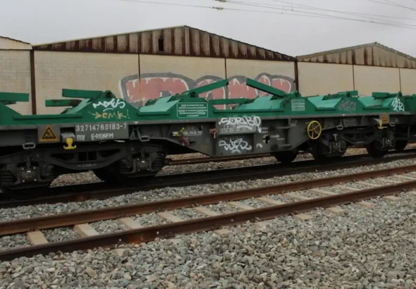 Un vagón ferroviario de color verde y gris circula por una vía, es una plataforma diseñada para llevar bobinas de acero al descubierto.