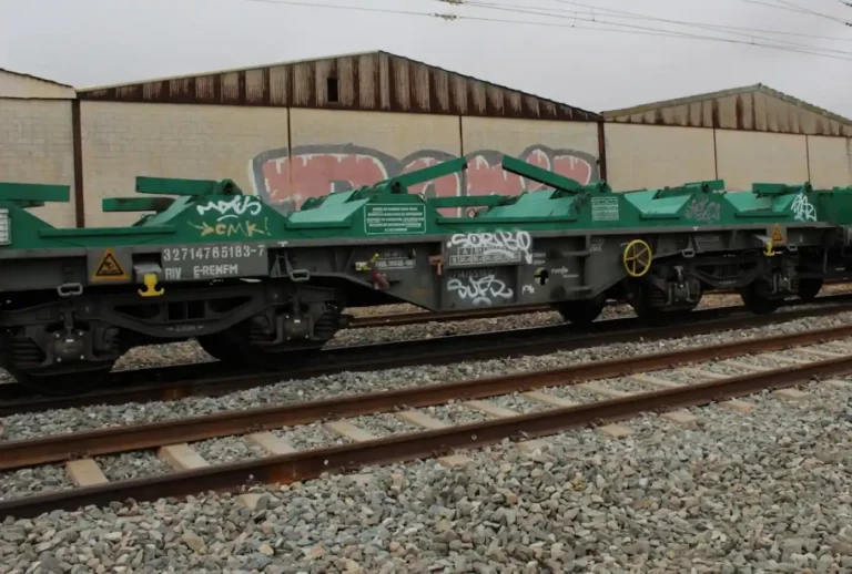 Un vagón ferroviario de color verde y gris circula por una vía, es una plataforma diseñada para llevar bobinas de acero al descubierto.