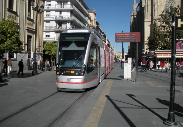 Un tranvía moderno con el número 301 y el logo de TUSSAM circula por una calle peatonal flanqueada por edificios históricos, árboles de naranja y una catedral gótica bajo un cielo despejado.