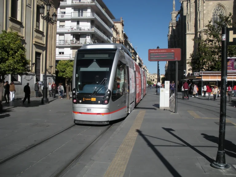 Un tranvía moderno con el número 301 y el logo de TUSSAM circula por una calle peatonal flanqueada por edificios históricos, árboles de naranja y una catedral gótica bajo un cielo despejado.