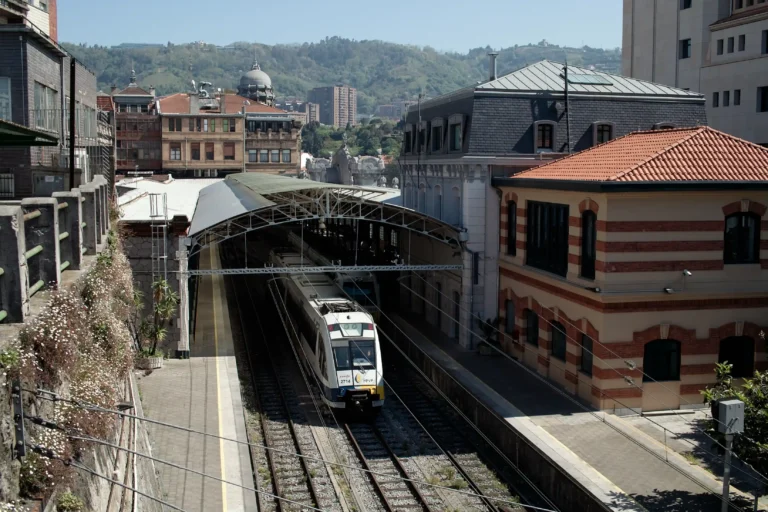Vista aérea de un tren blanco y azul de Renfe saliendo de una estación con cubierta metálica curva en Bilbao. A la derecha destaca un edificio histórico de ladrillo y piedra, y al fondo se ven las colinas verdes que rodean la ciudad bajo un cielo despejado.