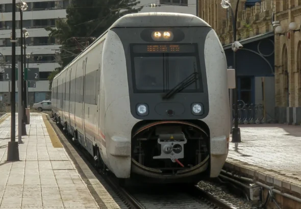 Vista frontal de un tren eléctrico blanco de la serie 449 de Renfe detenido en el andén de la antigua estación de trenes de Huelva Término, con el edificio histórico de estilo neomudéjar a la derecha y bloques de viviendas al fondo.
