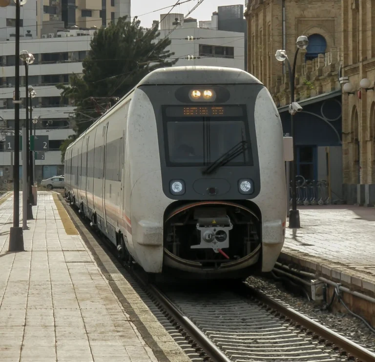 Vista frontal de un tren eléctrico blanco de la serie 449 de Renfe detenido en el andén de la antigua estación de trenes de Huelva Término, con el edificio histórico de estilo neomudéjar a la derecha y bloques de viviendas al fondo.