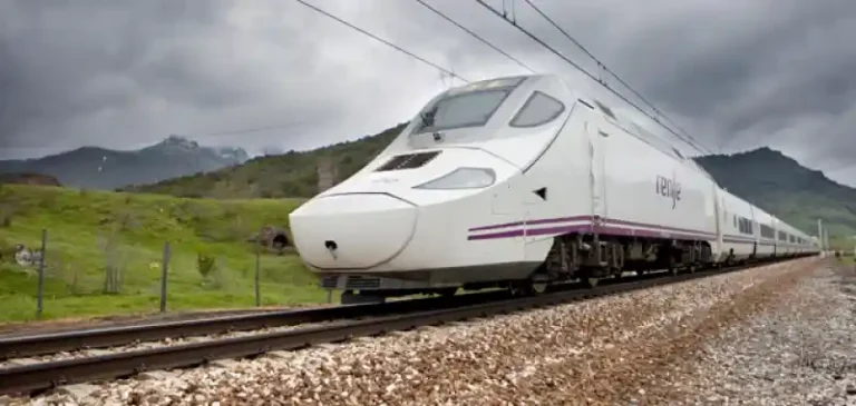 Tren Alvia de Renfe de color blanco y morado circulando por una vía de ferrocarril tradicional rodeada de vegetación y montañas bajo un cielo nublado.