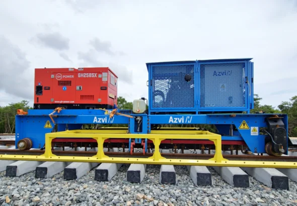 Maquinaria ferroviaria azul y amarilla de Azvi, equipada con un generador rojo, posicionada sobre traviesas de hormigón y balasto durante las fases de montaje y alineación automatizada de una vía de tren.
