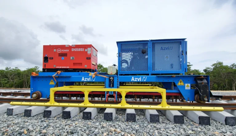 Maquinaria ferroviaria azul y amarilla de Azvi, equipada con un generador rojo, posicionada sobre traviesas de hormigón y balasto durante las fases de montaje y alineación automatizada de una vía de tren.