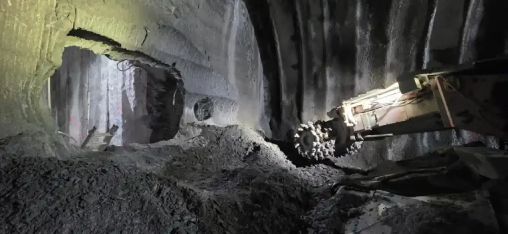 Fotografía del interior de un túnel del Metro de San Sebastián en construcción en mina. A la derecha, una máquina rozadora con su cabezal dentado se sitúa frente a una montaña de escombros de roca y tierra. A la izquierda, se observa una apertura o cale circular en la roca que conecta con otra sección del túnel excavado, donde hay una pequeña escalera apoyada.