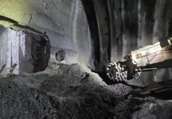 Fotografía del interior de un túnel del Metro de San Sebastián en construcción en mina. A la derecha, una máquina rozadora con su cabezal dentado se sitúa frente a una montaña de escombros de roca y tierra. A la izquierda, se observa una apertura o cale circular en la roca que conecta con otra sección del túnel excavado, donde hay una pequeña escalera apoyada.