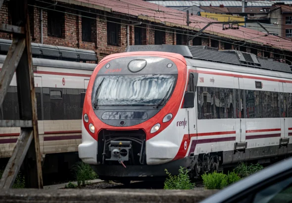 Primer plano de la cabina y frontal de un tren de Cercanías Renfe modelo Civia (unidad 219M) pintado de blanco y rojo, estacionado en una zona de vías de Bilbao frente a un edificio industrial de ladrillo visto.