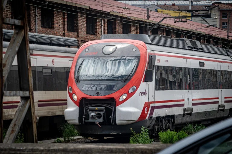 Primer plano de la cabina y frontal de un tren de Cercanías Renfe modelo Civia (unidad 219M) pintado de blanco y rojo, estacionado en una zona de vías de Bilbao frente a un edificio industrial de ladrillo visto.