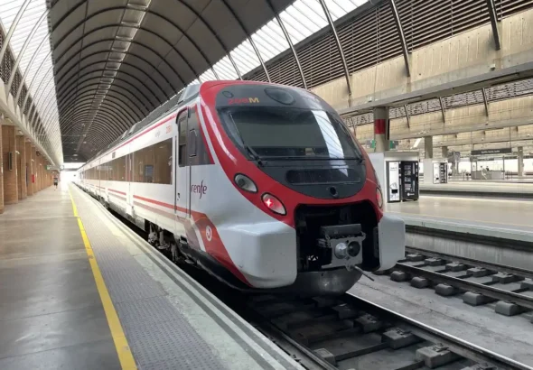 Un tren de Cercanías Renfe de color blanco y rojo estacionado junto a la vía bajo la nave acristalada de una estación ferroviaria.