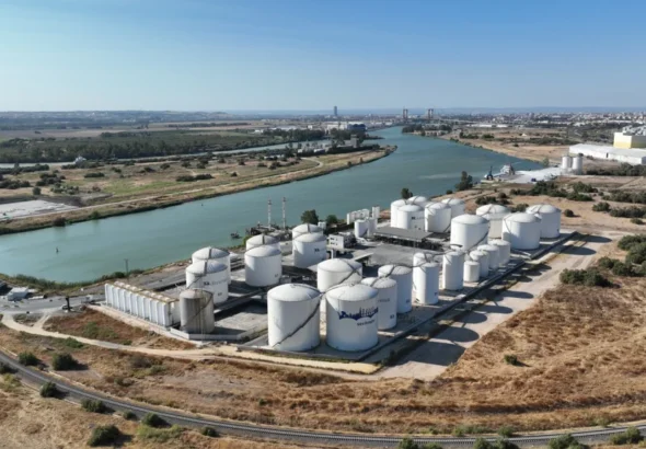 Vista aérea de un complejo industrial con grandes tanques cilíndricos blancos de almacenamiento de líquidos junto a un canal navegable y una vía de tren en el Puerto de Sevilla.