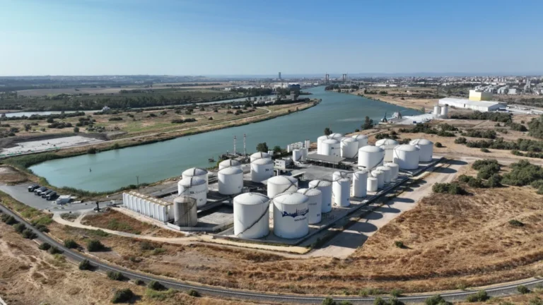 Vista aérea de un complejo industrial con grandes tanques cilíndricos blancos de almacenamiento de líquidos junto a un canal navegable y una vía de tren en el Puerto de Sevilla.