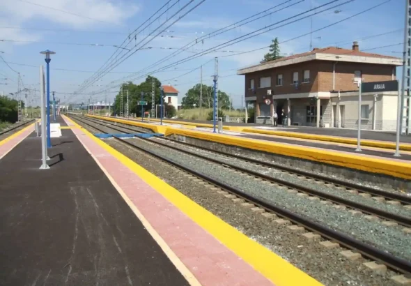 Vías del tren y andenes de la estación de ferrocarril de Araia, con el edificio principal de ladrillo a la derecha y catenarias sobre las vías.