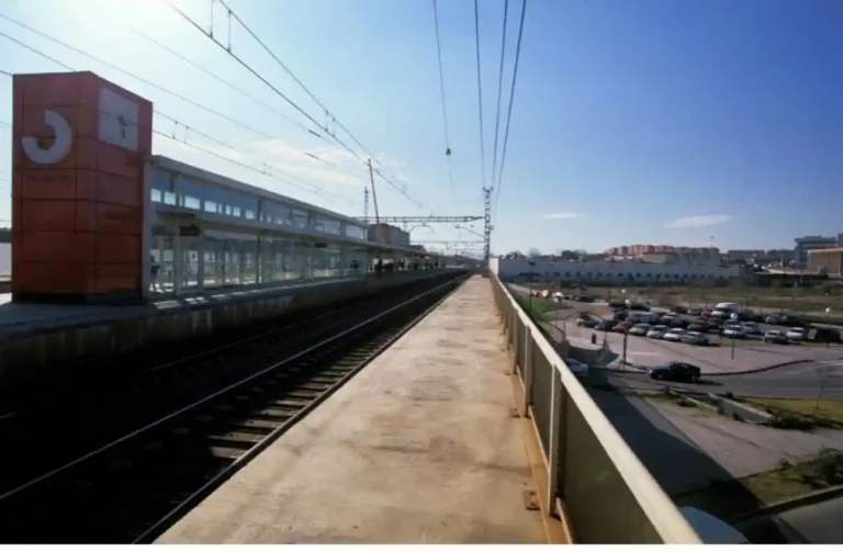 Vista panorámica del andén y las vías de la estación de Cercanías de Vallecas en Madrid, con la estructura metálica naranja y el reloj en primer plano.