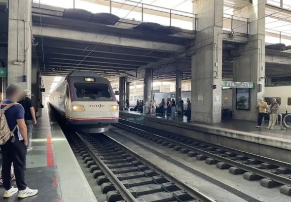 Un tren de pasajeros de Renfe de color blanco y morado entra a la estación de Córdoba Julio Anguita, mientras varios viajeros esperan en los andenes cubiertos de hormigón.
