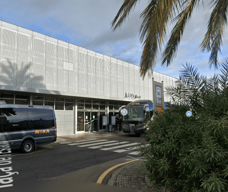 Vista exterior de la estación ferroviaria de Reus con un autobús y una furgoneta de la compañía Nika estacionados frente a la fachada moderna de paneles metálicos perforados.