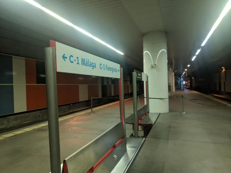 Andén subterráneo de la estación de Victoria Kent con bancos metálicos y un cartel indicador de la línea C-1 hacia Málaga y Fuengirola, bajo una hilera de luces de techo encendidas.
