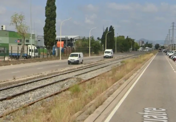 Fotografía a pie de calle de las vías del tren que dividen las calzadas de la calle 4 en la Zona Franca de Barcelona. Se observa el balasto con vegetación seca en los bordes, furgonetas circulando por la carretera de la izquierda, coches aparcados a la derecha y naves industriales al fondo bajo un cielo parcialmente nublado.