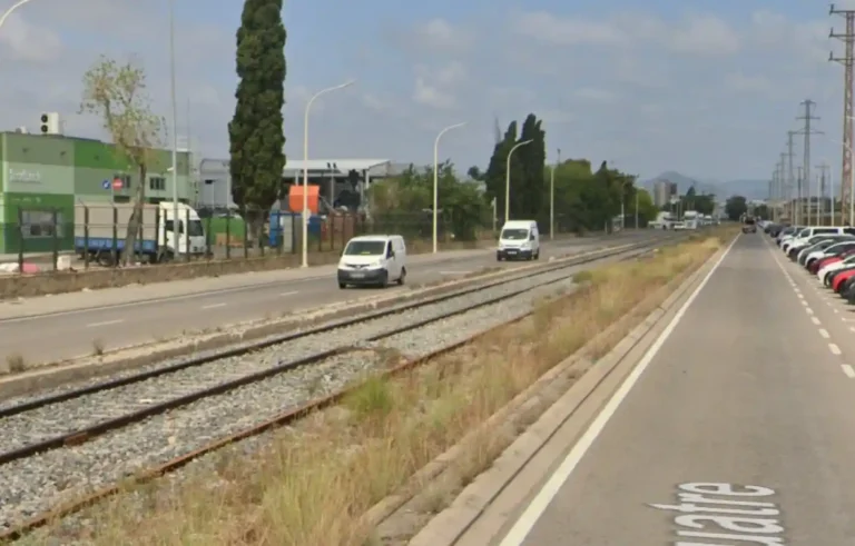 Fotografía a pie de calle de las vías del tren que dividen las calzadas de la calle 4 en la Zona Franca de Barcelona. Se observa el balasto con vegetación seca en los bordes, furgonetas circulando por la carretera de la izquierda, coches aparcados a la derecha y naves industriales al fondo bajo un cielo parcialmente nublado.