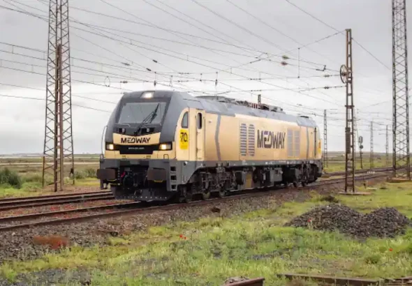 Una locomotora de tracción diésel de color dorado y gris de la compañía Medway circulando de frente por una vía de tren bajo un tendido de catenarias y postes eléctricos en un día nublado.