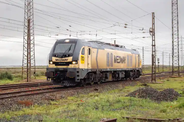 Una locomotora de tracción diésel de color dorado y gris de la compañía Medway circulando de frente por una vía de tren bajo un tendido de catenarias y postes eléctricos en un día nublado.