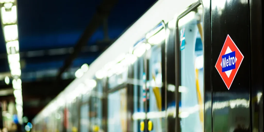 Plano de detalle del lateral negro de un tren del Metro de Madrid, mostrando el logotipo oficial en forma de rombo rojo, blanco y azul, con el interior del coche iluminado y el andén desenfocados al fondo.