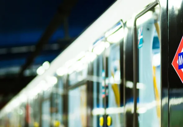 Plano de detalle del lateral negro de un tren del Metro de Madrid, mostrando el logotipo oficial en forma de rombo rojo, blanco y azul, con el interior del coche iluminado y el andén desenfocados al fondo.
