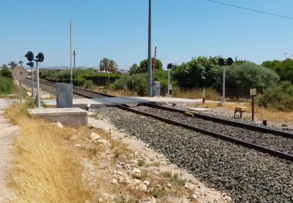 Vista en perspectiva de un paso a nivel ferroviario de vía única en una zona rural y despejada de Elche. Se observa la vía del tren cruzando un camino vecinal asfaltado, equipada con señales de Stop, semáforos de advertencia luminosa con pantallas negras circulares, una barrera levantada con franjas rojas y blancas, y cartelería técnica. Al fondo se aprecian palmeras y vegetación mediterránea bajo un cielo despejado.