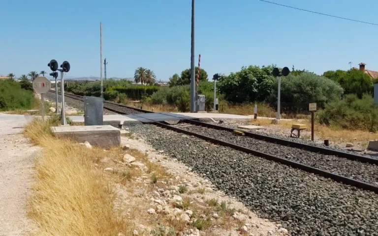 Vista en perspectiva de un paso a nivel ferroviario de vía única en una zona rural y despejada de Elche. Se observa la vía del tren cruzando un camino vecinal asfaltado, equipada con señales de Stop, semáforos de advertencia luminosa con pantallas negras circulares, una barrera levantada con franjas rojas y blancas, y cartelería técnica. Al fondo se aprecian palmeras y vegetación mediterránea bajo un cielo despejado.