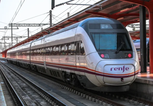 Tren Avant de la serie 121 de Renfe de color blanco y morado detenido en el andén de una estación ferroviaria bajo una marquesina roja, mostrando las vías y la catenaria.