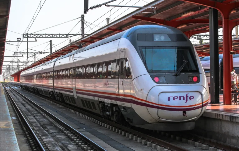 Tren Avant de la serie 121 de Renfe de color blanco y morado detenido en el andén de una estación ferroviaria bajo una marquesina roja, mostrando las vías y la catenaria.