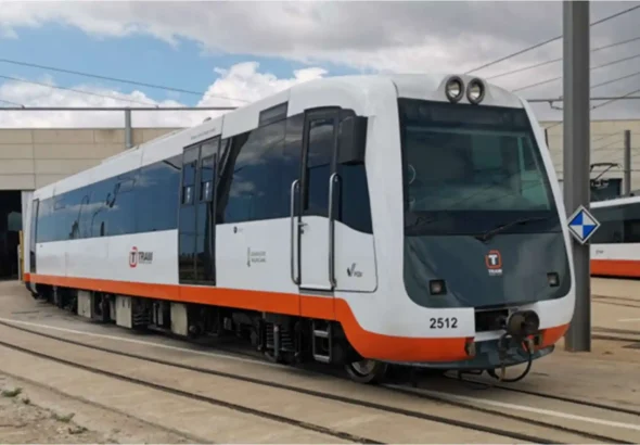 Tren de pasajeros de color blanco y gris con una franja naranja inferior, rotulado con el logotipo de TRAM y el número 2512, detenido en las vías exteriores de unas instalaciones ferroviarias bajo un cielo parcialmente nublado.