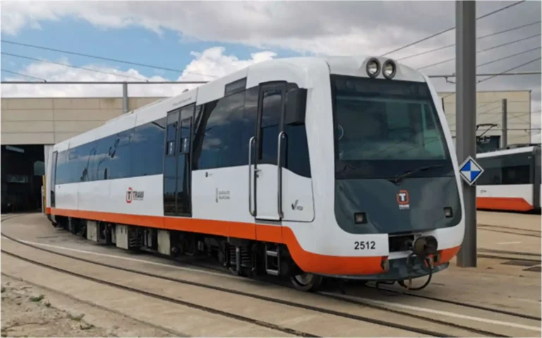 Tren de pasajeros de color blanco y gris con una franja naranja inferior, rotulado con el logotipo de TRAM y el número 2512, detenido en las vías exteriores de unas instalaciones ferroviarias bajo un cielo parcialmente nublado.