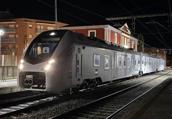 Fotografía nocturna de un tren moderno de color blanco y gris de la serie 312 de Ferrocarrils de la Generalitat de Catalunya (FGC) estacionado en las vías de una estación. En el lateral del vagón se lee el texto "Vehicle en proves" y el logotipo de Stadler. Al fondo se observa un edificio clásico iluminado.