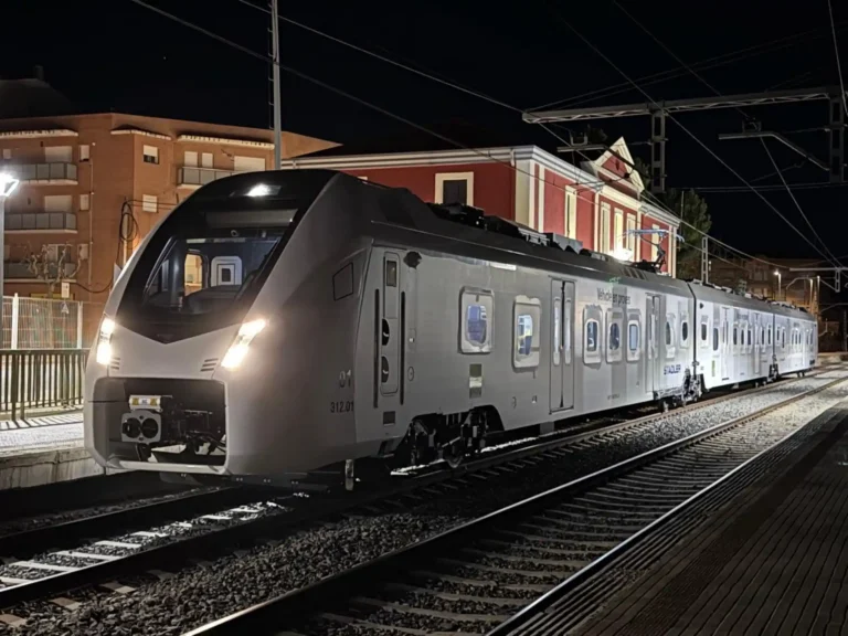 Fotografía nocturna de un tren moderno de color blanco y gris de la serie 312 de Ferrocarrils de la Generalitat de Catalunya (FGC) estacionado en las vías de una estación. En el lateral del vagón se lee el texto "Vehicle en proves" y el logotipo de Stadler. Al fondo se observa un edificio clásico iluminado.