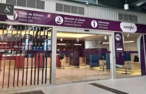 Fachada acristalada de la oficina de Renfe en la estación de tren de Santiago de Compostela, con cartelería morada que indica las zonas de venta de billetes, atención al cliente e información.