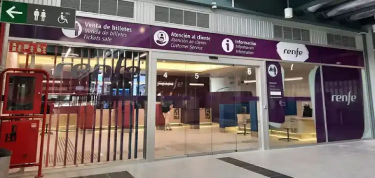 Fachada acristalada de la oficina de Renfe en la estación de tren de Santiago de Compostela, con cartelería morada que indica las zonas de venta de billetes, atención al cliente e información.