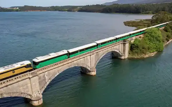 Vista aérea del tren turístico Costa Verde Express cruzando un viaducto sobre un estuario rodeado de vegetación, durante las pruebas con combustible 100% renovable HVO.Vista aérea del tren turístico Costa Verde Express cruzando un viaducto sobre un estuario rodeado de vegetación, durante las pruebas con combustible 100% renovable HVO.