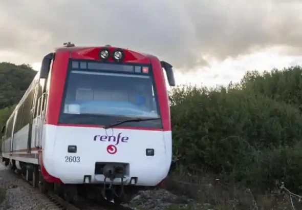 Un tren de ancho métrico de Renfe, de la serie 2600, pintado de blanco y rojo con el número de unidad 2603, avanza por una vía única rodeada de vegetación densa y árboles bajo un cielo nublado.