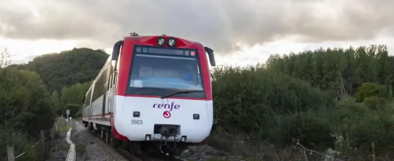 Un tren de ancho métrico de Renfe, de la serie 2600, pintado de blanco y rojo con el número de unidad 2603, avanza por una vía única rodeada de vegetación densa y árboles bajo un cielo nublado.