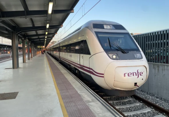 Tren de media/larga distancia de Renfe estacionado en la vía 1 del andén de la estación de tren de Huelva bajo una marquesina.