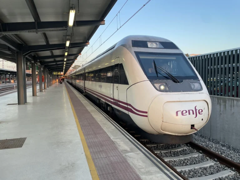 Tren de media/larga distancia de Renfe estacionado en la vía 1 del andén de la estación de tren de Huelva bajo una marquesina.