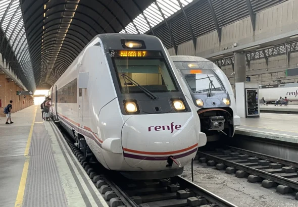 Tren de Media Distancia de Renfe de color blanco con franja morada estacionado en la vía del andén número 12 de una estación cubierta, junto a otro convoy.