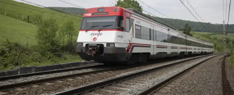 Un tren de Cercanías Renfe de color blanco y rojo circula por una vía de tren rodeada de prados verdes y colinas en el territorio de Gipuzkoa, dirección andonain.