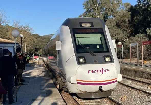 Tren blanco y morado de Renfe estacionado en la vía junto al andén de la estación de Cazalla-Constantina en un día despejado, con pasajeros esperando en el lateral y árboles de fondo.
