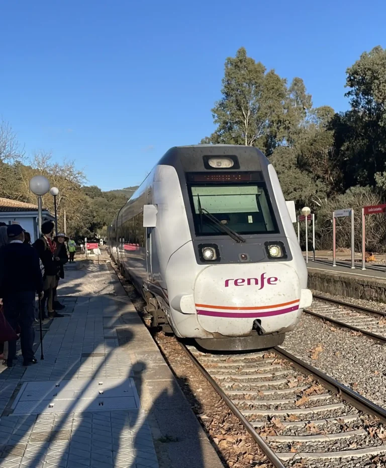Tren blanco y morado de Renfe estacionado en la vía junto al andén de la estación de Cazalla-Constantina en un día despejado, con pasajeros esperando en el lateral y árboles de fondo.