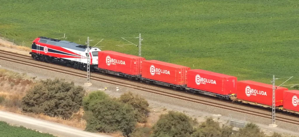 Vista aérea de un tren de mercancías con locomotora roja, blanca y azul arrastrando varios vagones con contenedores rojos de la empresa Boluda Lines a través del campo.